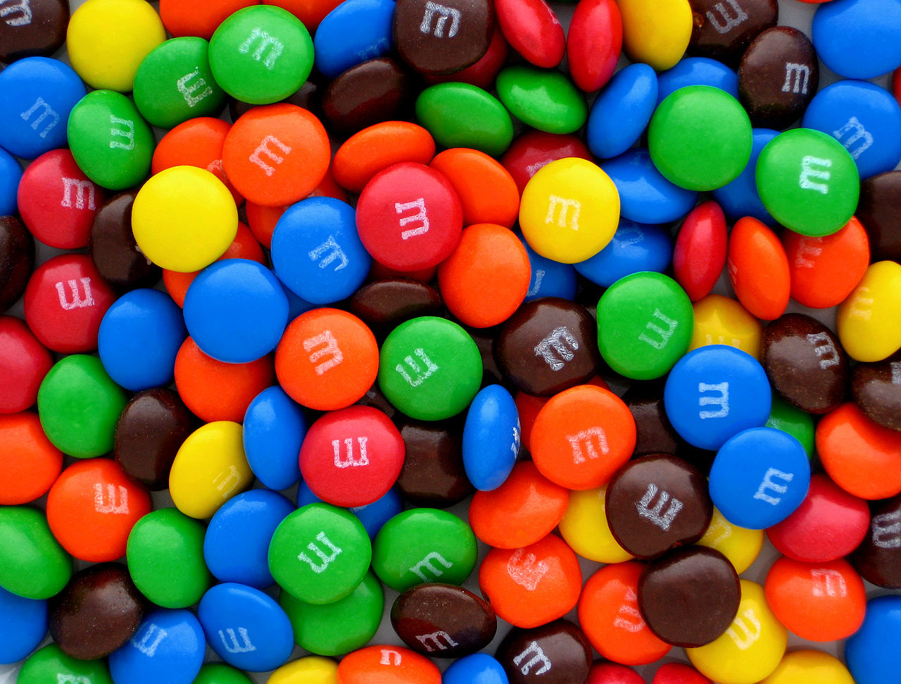 A close-up photo of a pile of M&M candies in assorted colours: red, blue, yellow, green, orange, and brown, each a small glossy ellipsoid with a white letter M printed on one side